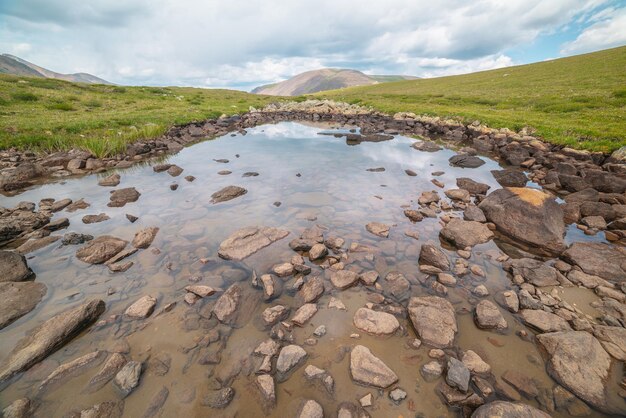 Pintoresco paisaje alpino con un pequeño lago de montaña en una colina cubierta de hierba verde bajo un cielo nublado Espectacular vista de montaña a un pequeño lago con reflejo de nubes Agua transparente en un lago alpino con fondo pedregoso