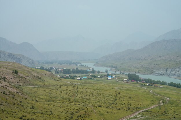 Pintoresco paisaje alpino con pueblo de montaña por río de montaña en la niebla. Paisaje brumoso atmosférico con campo en la orilla del río. Hermosa vista al pueblo de montaña a orillas del río cuando llueve.