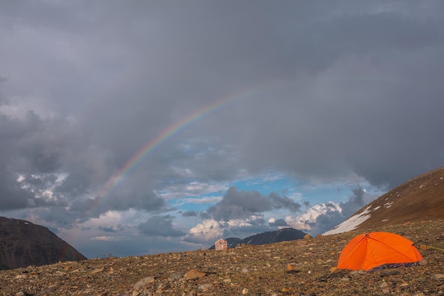 Pintoresco paisaje de montaña con carpa naranja vívida a la luz del sol y arco iris en cielo nublado Hermoso paisaje alpino con carpa bajo arco iris en nublado Carpa naranja y arco iris alto en clima lluvioso