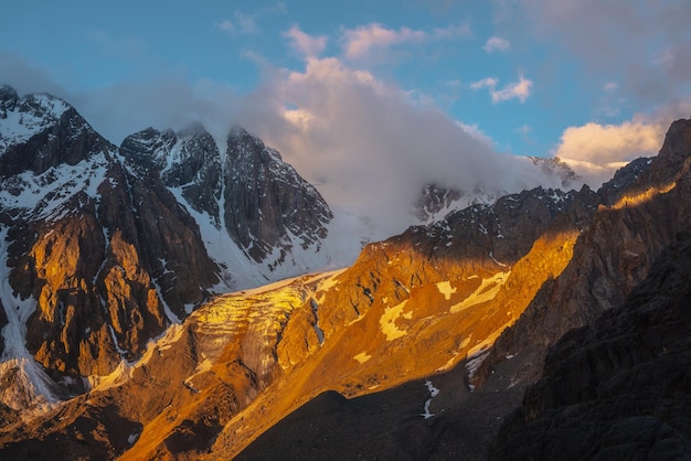 Pintoresco paisaje de montaña con cima de montaña nevada y glaciar en nubes bajas en colores dorados del amanecer Vista colorida de la montaña al glaciar vertical con cascada de hielo en la luz del sol dorada de la mañana en nubes bajas