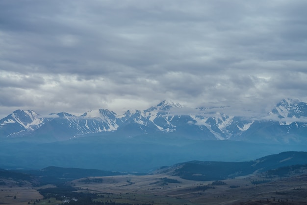 Pintoresco paisaje de montaña con gran cordillera nevada entre nubes bajas y bosque verde en el valle temprano en la mañana. Paisaje alpino atmosférico con cresta de alta montaña blanca azul bajo un cielo nublado.