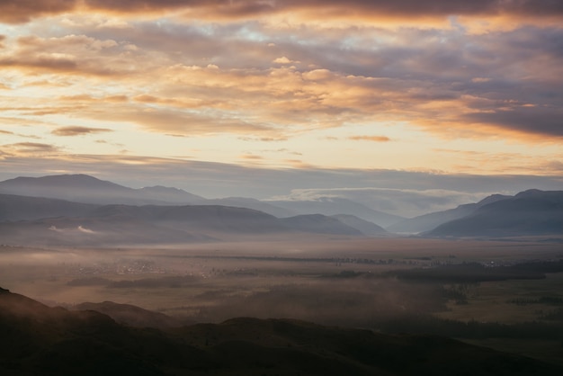 Pintoresco paisaje de montaña con nubes doradas por encima de la aldea entre siluetas de montañas bajo el cielo nublado del amanecer. El paisaje alpino atmosférico del campo en nubes bajas en el color que ilumina la puesta del sol.