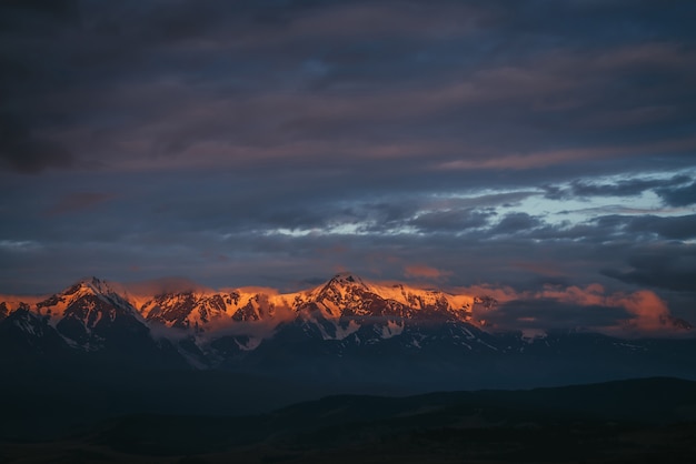 Pintoresco paisaje montañoso con gran cordillera nevada iluminada por el sol del amanecer entre nubes bajas. Impresionante paisaje alpino con crestas de alta montaña al atardecer o al amanecer. Gran glaciar en la parte superior en luz naranja.