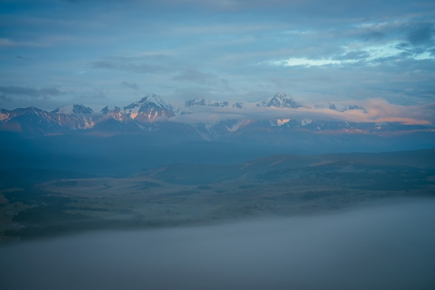 Pintoresco paisaje montañoso con una gran cordillera nevada iluminada por el sol anaranjado del amanecer sobre una densa niebla entre las nubes bajas en el crepúsculo. Impresionante paisaje de la mañana con cresta de alta montaña bajo un cielo nublado azul.