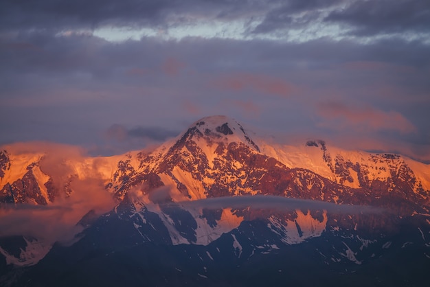 Pintoresco paisaje montañoso con grandes montañas nevadas iluminadas por el sol del amanecer entre nubes bajas. Impresionante paisaje alpino con pináculo de alta montaña al atardecer o al amanecer. Gran glaciar en la parte superior en luz naranja.