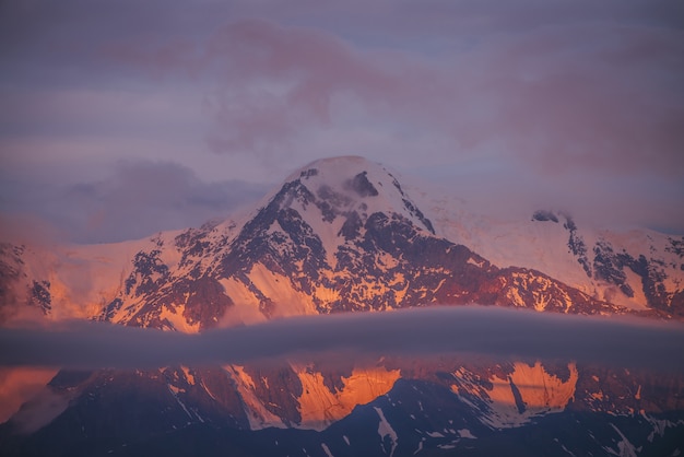 Pintoresco paisaje montañoso con grandes montañas nevadas iluminadas por el sol del amanecer entre nubes bajas.