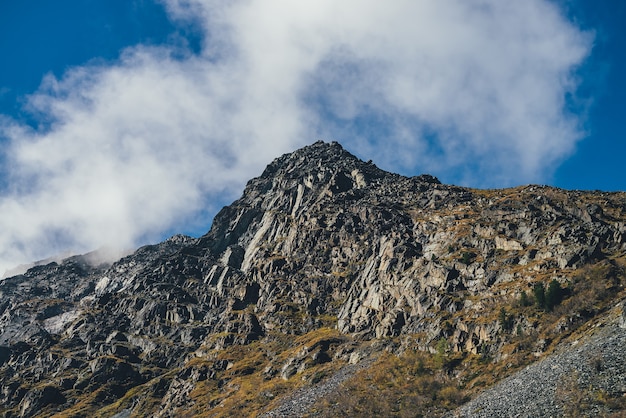 Pintoresco paisaje soleado de otoño con alta montaña rocosa con cima puntiaguda en nubes bajas bajo un cielo azul. Impresionante vista alpina al hermoso pináculo agudo bajo el sol. Hermoso pico de la montaña en la luz del sol.