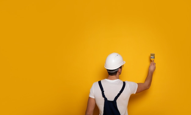 Pintura amarilla. Foto de primer plano del hombre con traje de trabajo y casco blanco, que está pintando una pared en amarillo.