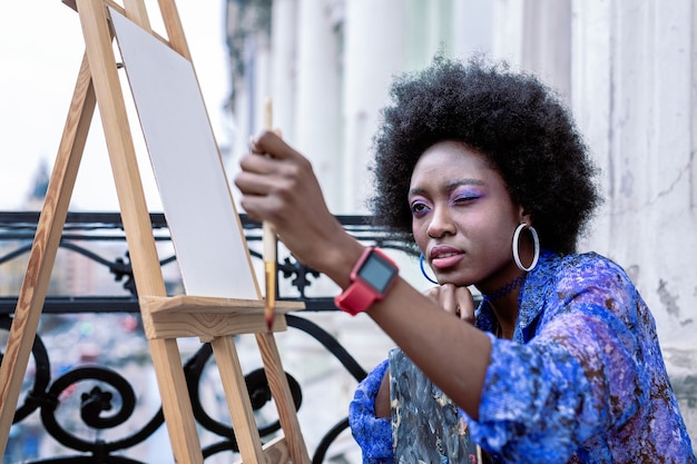 Pintura exterior. Artista elegante y talentoso con maquillaje brillante y vistiendo un vestido azul pintando afuera