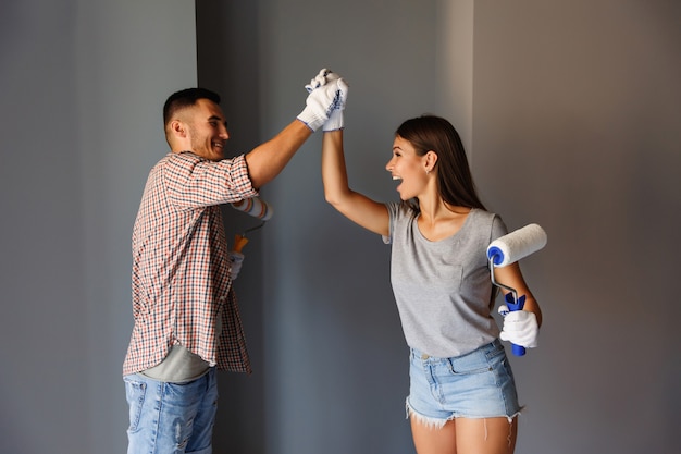 Pintura final en la casa nueva. Feliz pareja celebrando final de pintura con rodillo de pintura en la pared gris