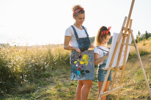 Pintura de la madre de la belleza con su pequeña hija. Mujer elegante dibujando la imagen con niña. Niño lindo con una camiseta blanca y pantalones vaqueros azules.