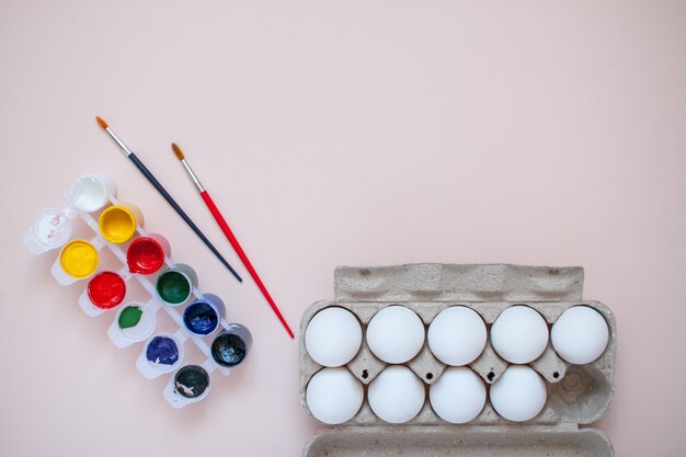 Pinturas brillantes y dos pinceles sobre una mesa de color rosa claro. Casete de huevos. Preparándose para pintar huevos para