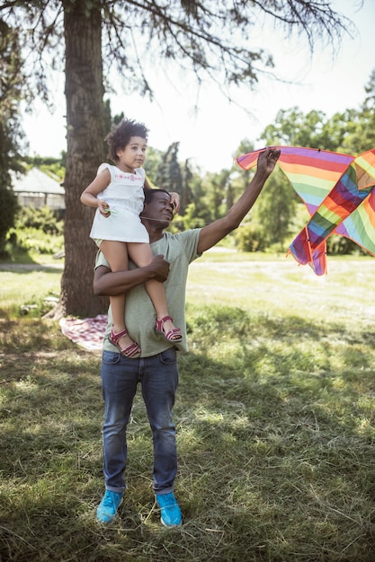 Foto pipa. homem de pele escura mostrando uma pipa para a filha sentada em seus ombros