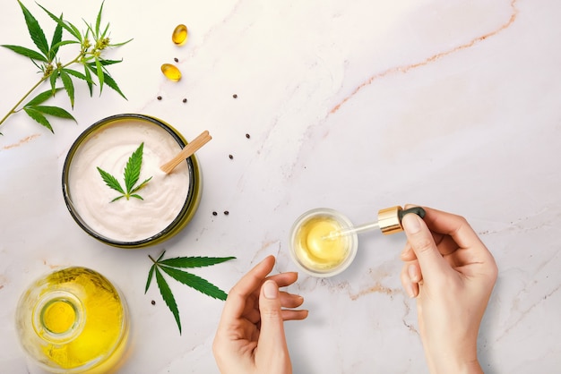 Pipetar com óleo cosmético CBD em mãos femininas com cosméticos, creme com cannabis e folhas de cânhamo