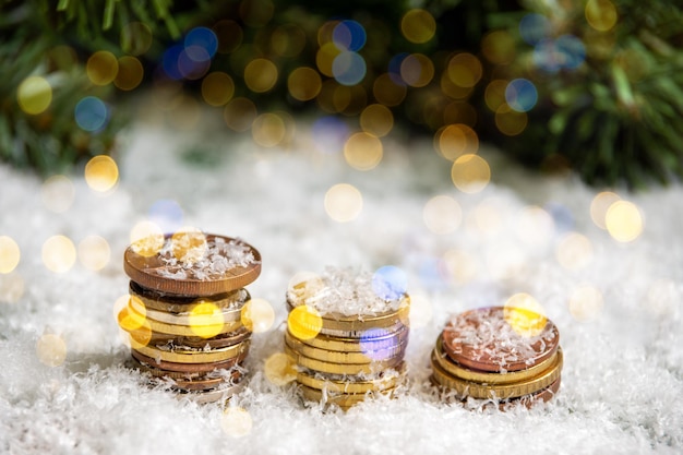 Pirámides de oro de monedas cubiertas de nieve sobre un fondo de ramas de abeto verde con bokeh de una guirnalda. Concepto de riqueza en el nuevo año.