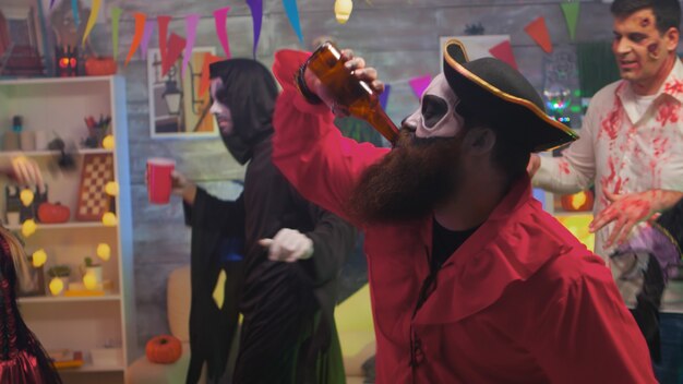 Pirata guapo con un hacha bebiendo cerveza celebrando halloween con sus amigos asustadizos bailando en la habitación decorada.