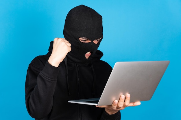 Pirata informático con máscara de esquí negra celebrando un nuevo delito cibernético mientras sostiene una computadora portátil