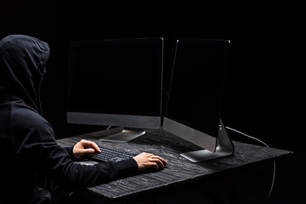 Pirata informático usando el mouse de la computadora y mirando monitores de computadora con pantalla en blanco aislada en negro