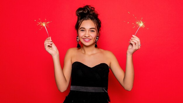 Pirotecnia y concepto de personas mujer joven sonriente o adolescente mujer feliz con bengalas celebrar en vestido negro sobre fondo rojo niña sonriendo a la cámara