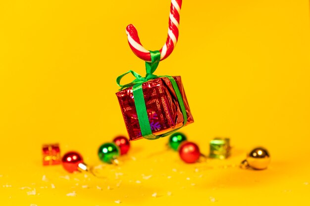 Piruleta de bastón de caramelo sosteniendo una caja de regalo con un regalo de Navidad sobre un fondo amarillo, dulces navideños con adornos de año nuevo.