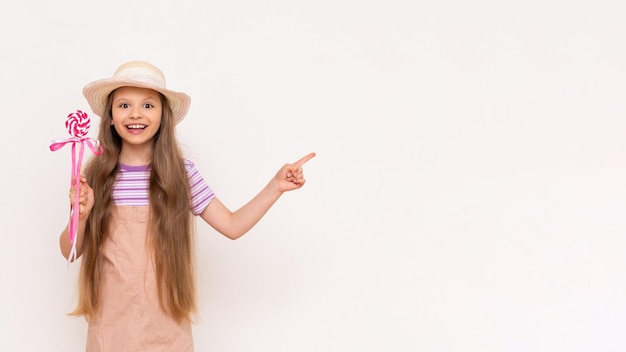 Una piruleta de caramelo en manos de una hermosa niña Un bebé con un vestido de verano y un sombrero de verano señala un fondo blanco aislado con su dedo índice