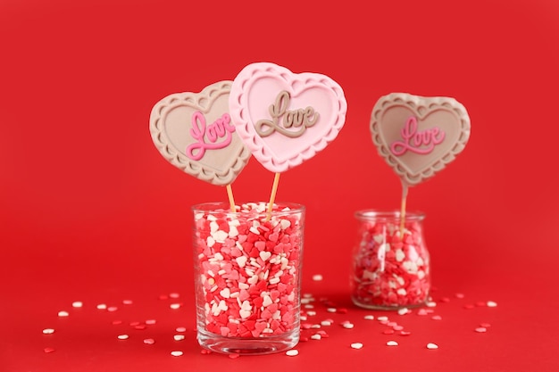 Pirulitos em forma de coração feitos de chocolate com granulado em copos sobre fundo vermelho