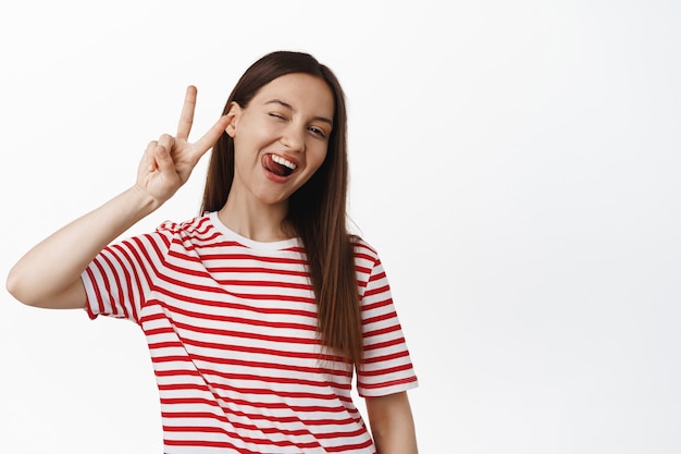 Piscadela de mulher jovem feliz, mostra a língua boba e o símbolo de V da paz, humor positivo de verão, vestindo uma camiseta vermelha listrada, em pé contra uma parede branca.