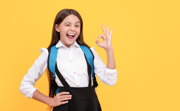 Piscando a criança feliz em uniforme escolar carrega mochila mostrando o aluno excelente gesto ok