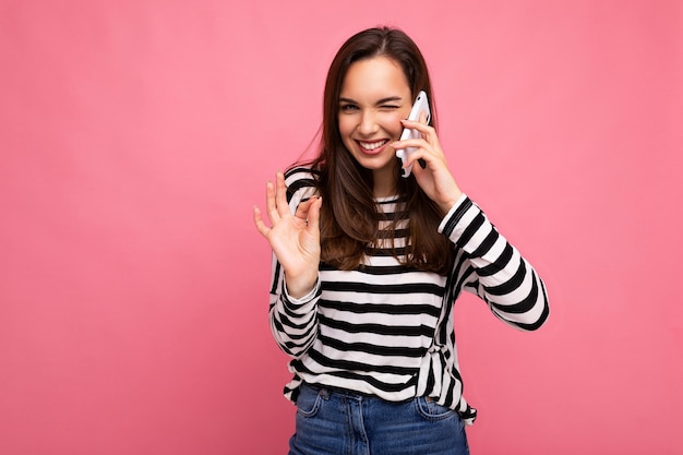 Piscando Mulher jovem muito feliz falando ao telefone, vestindo uma blusa listrada isolada sobre um fundo com espaço de cópia, mostrando o gesto de okey, olhando para a câmera.