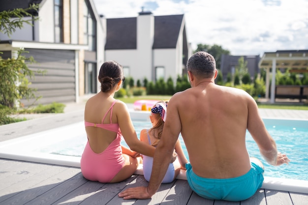 Piscina cerca de la casa. Familia feliz vistiendo trajes de baño sentado junto a la piscina cerca de su casa privada