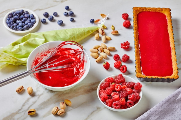 Pistazien-Ganache-Tarte mit Himbeer-Pektin-Gelee serviert auf einer Marmoroberfläche mit Schneebesen, Spritzbeutel gefüllt mit weißer Schokolade und Pistazien-Ganache, frischen Blaubeeren und Himbeeren, Ansicht von oben