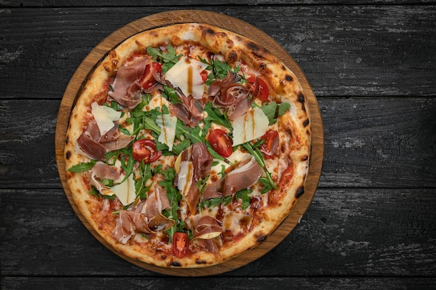 Pizza con carne parmesano y tomates en una tabla de madera sobre una mesa oscura