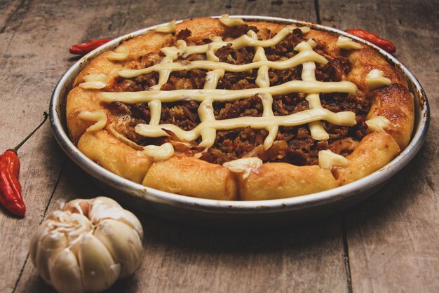 Pizza con carne de res y queso crema rodeada de chiles y ajo servida en una mesa de madera