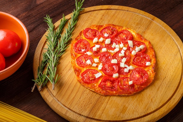 Foto pizza casera con tomates y mozzarella en una tabla redonda de madera junto a una ramita de romero y tomates