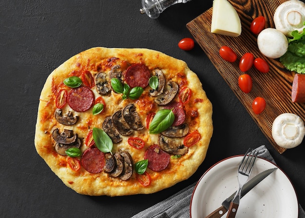 Foto pizza con champiñones salami y albahaca servida con plato en mesa oscura con vista superior de ingredientes