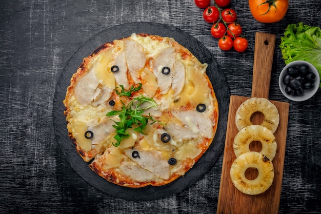 Pizza com frango, abacaxi, queijo e azeitonas na pedra e um fundo preto de madeira riscado