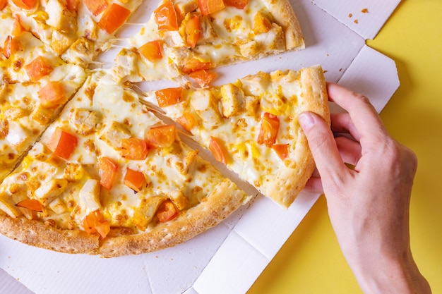 Pizza com frango e tomate em uma caixa. Mão tira uma fatia de pizza.