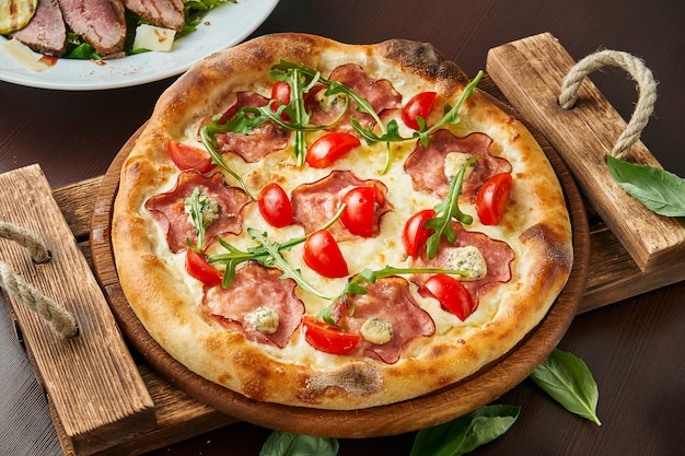 Pizza com presunto, queijo, tomate cereja, rúcula na bandeja de madeira.