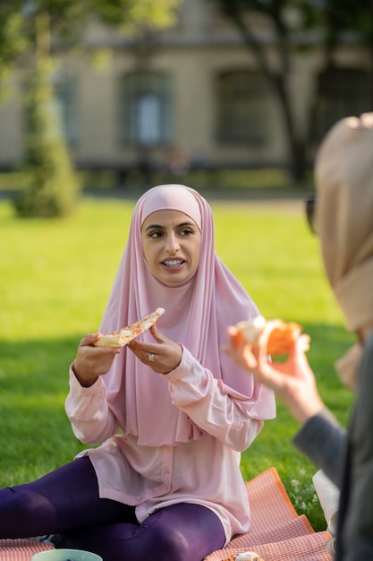 Pizza com um amigo. mulher muçulmana de olhos escuros usando um hijab rosa comendo pizza com uma amiga