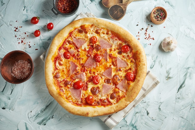 Pizza crocante recém-assada com tomate cereja, mussarela e presunto em uma mesa cinza em uma composição com ingredientes e utensílios de cozinha