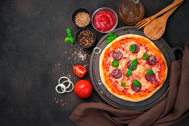 Pizza de calabresa em uma tábua de cortar marrom entre ingredientes e guardanapos em um fundo de concreto.