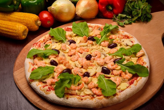 Pizza de camarão e cogumelos com rúcula em uma placa de madeira e legumes no fundo.