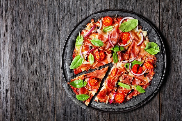 Pizza de massa preta com fatias de jamon, queijo mussarela, tomate, folhas de espinafre e cebola roxa em uma bandeja preta em uma mesa de madeira vista horizontal de cima