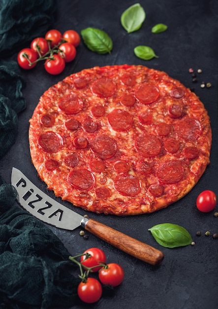 Pizza de pepperoni quente e picante fresca e redonda com faca e tomate com manjericão no fundo da mesa de cozinha preta