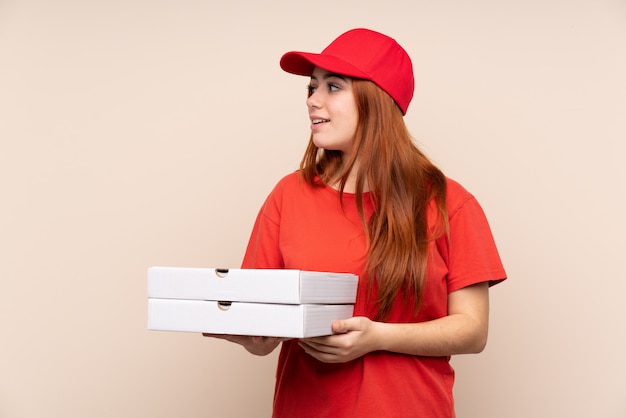 Pizza entrega mujer adolescente sosteniendo una pizza sobre pared aislada mirando hacia el lado