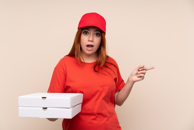 Pizza entrega mujer adolescente sosteniendo una pizza sobre pared aislada sorprendido y apuntando con el dedo hacia un lado