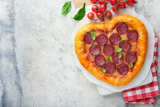 Pizza en forma de corazón del día de San Valentín con mozzarella pepperoni y botella de vino de albahaca caja de regalo de dos copas de vino sobre fondo gris claro Idea para una cena romántica Día de San Valentín Vista superior Maqueta