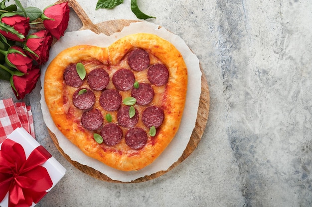 Pizza en forma de corazón del día de San Valentín con mozzarella pepperoni y botella de vino de albahaca caja de regalo de dos copas de vino sobre fondo gris claro Idea para una cena romántica Día de San Valentín Vista superior Maqueta