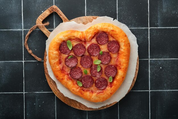 Pizza en forma de corazón del día de San Valentín con mozzarella pepperoni y botella de vino de albahaca caja de regalo de dos copas de vino sobre fondo negro Idea para una cena romántica Día de San Valentín Vista superior Maqueta