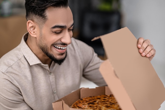 pizza fresca Primer plano de una cara sonriente contenta de un hombre joven con los ojos cerrados sosteniendo una caja de pizza abierta